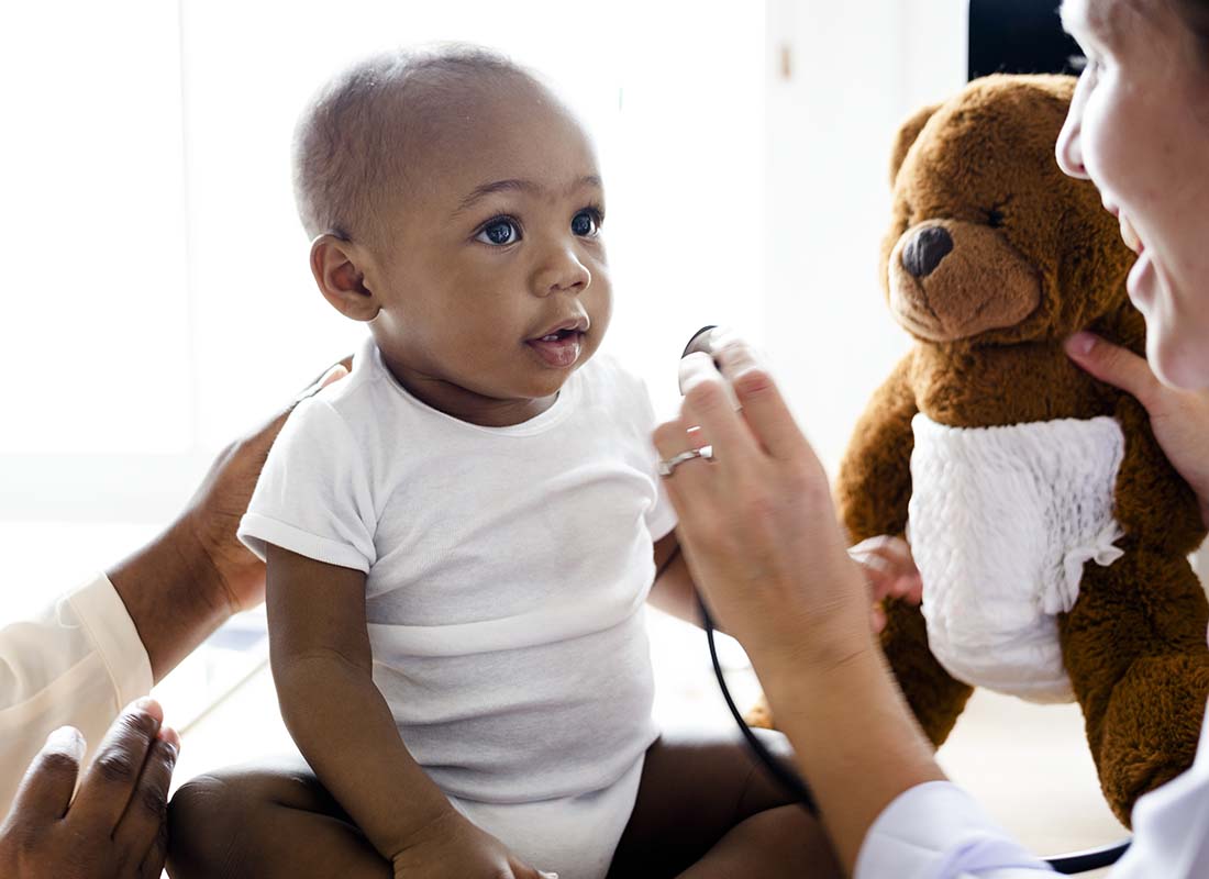 Individual Health Insurance - Baby during a Doctor Visit for a Wellness Checkup with the Doctor Holding a Child’s Teddy Bear to Try and Keep Child’s Attention and to Stay Positive