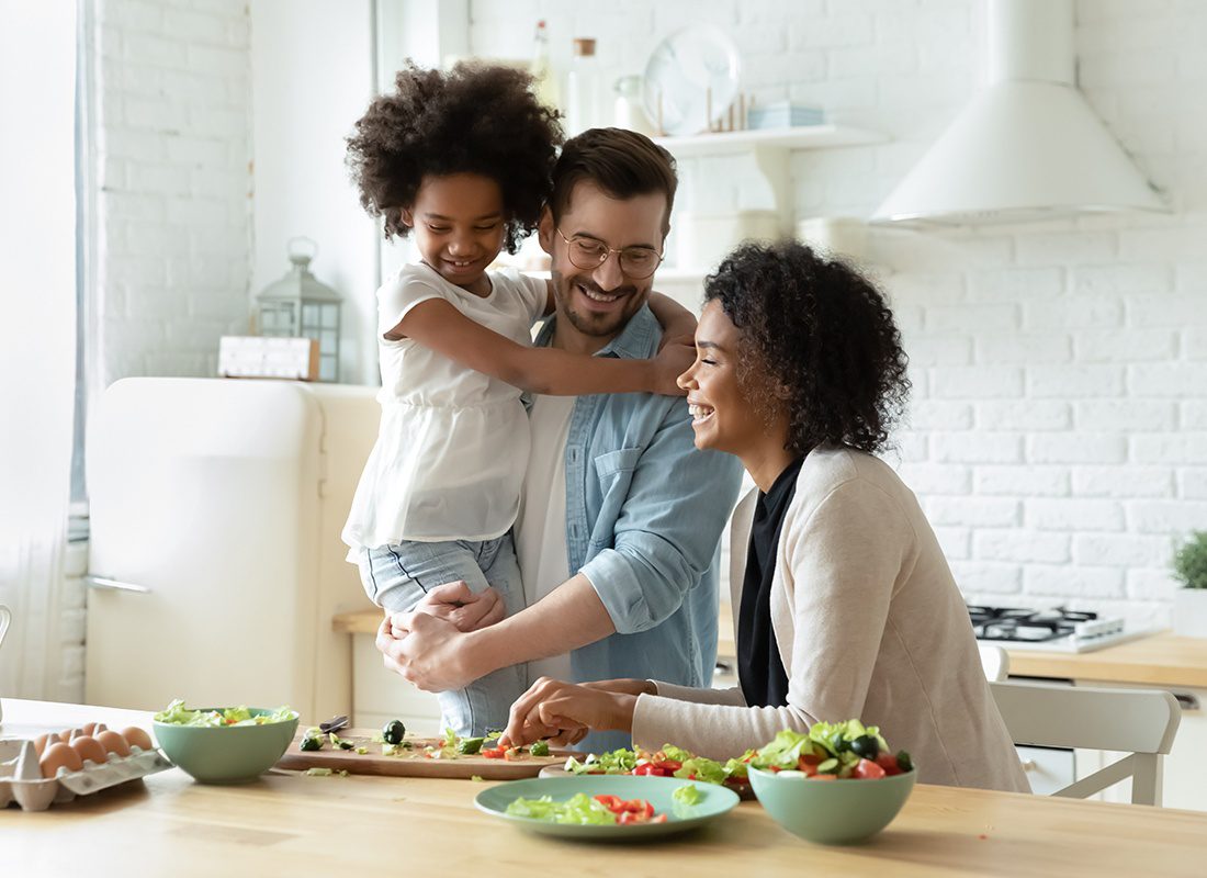Employee Benefits - Happy Father Holds Daughter in Arms While Mother Prepares Salad in the Kitchen